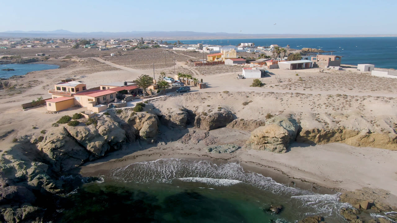 Aerial view in Bahia Ascuncion with surf reaching the lower beach, large rocks casting shadows near the cove, low coastal structures behind the sand, and no visible people.
