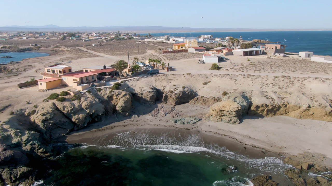 Wide aerial coastline with beach, rocks, town buildings, and ocean horizon