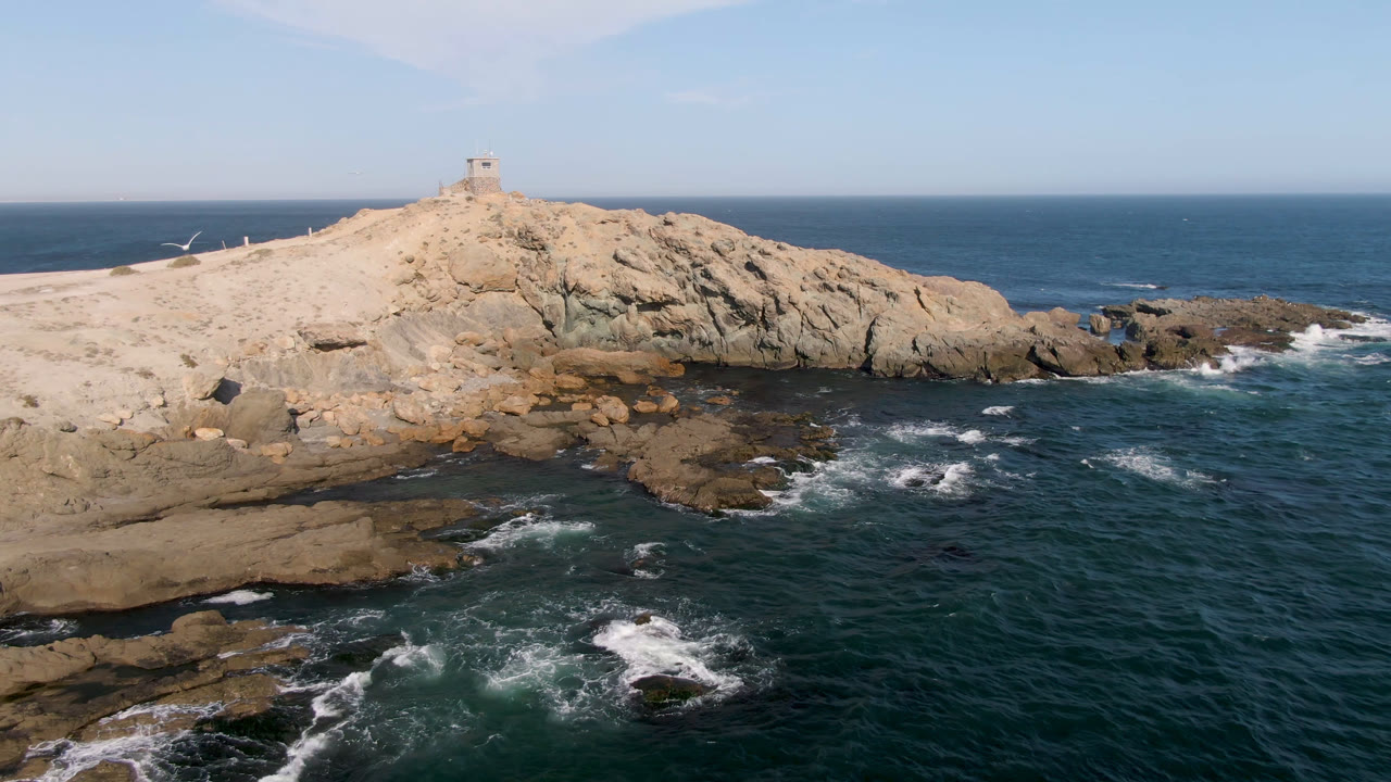 Aerial frame of Bahia Ascuncion, Baja California Sur, with dark ocean in front, white surf around rock shelves, a sandy headland, a small lighthouse on the ridge, and no visible people.