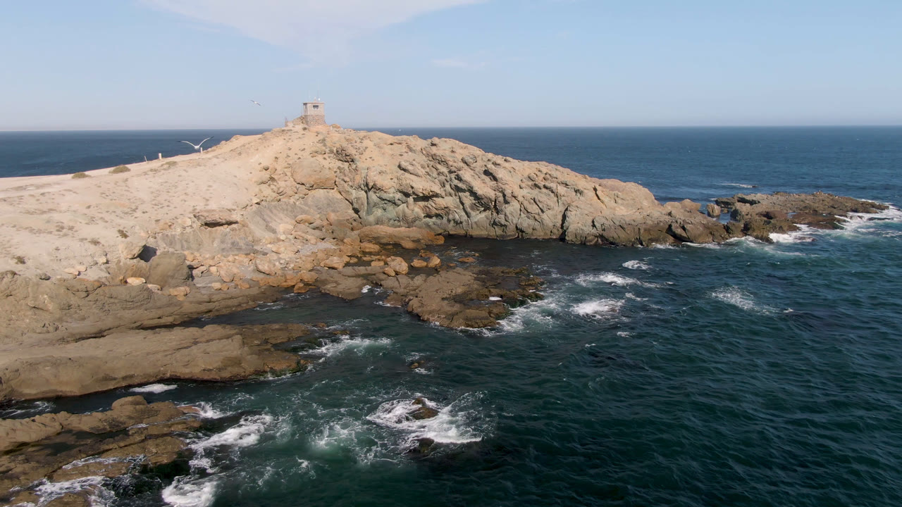 Wide aerial view in Bahia Ascuncion showing waves spreading across low rocks, tan sand leading up to a lighthouse, blue sea beyond, and no visible people.