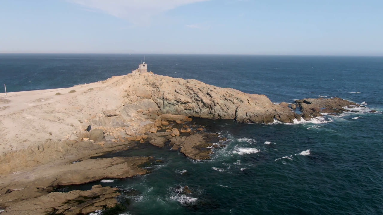 Aerial frame from Bahia Ascuncion showing a pale rocky headland, dark ocean water, white surf at the rocks, a small ridge structure, and no visible people.