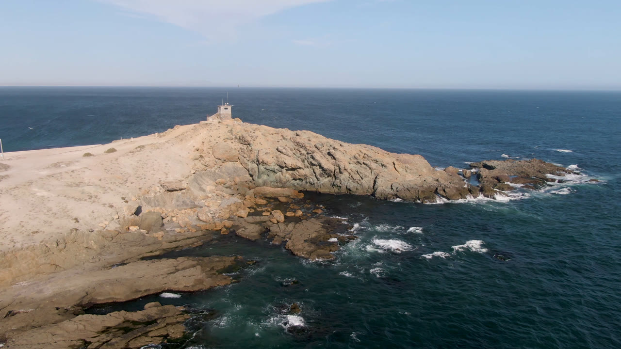 Aerial view over Bahia Ascuncion with the rocky point centered higher in frame, surf curling around exposed rocks, open water to the right, and no visible people.