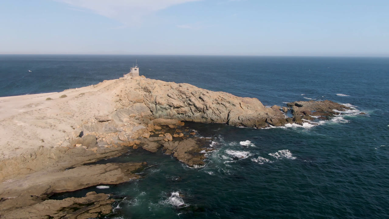 Aerial frame from Bahia Ascuncion showing the rocky cliff face, darker water close to shore, small whitewater patches, the ridge structure near the top, and no visible people.