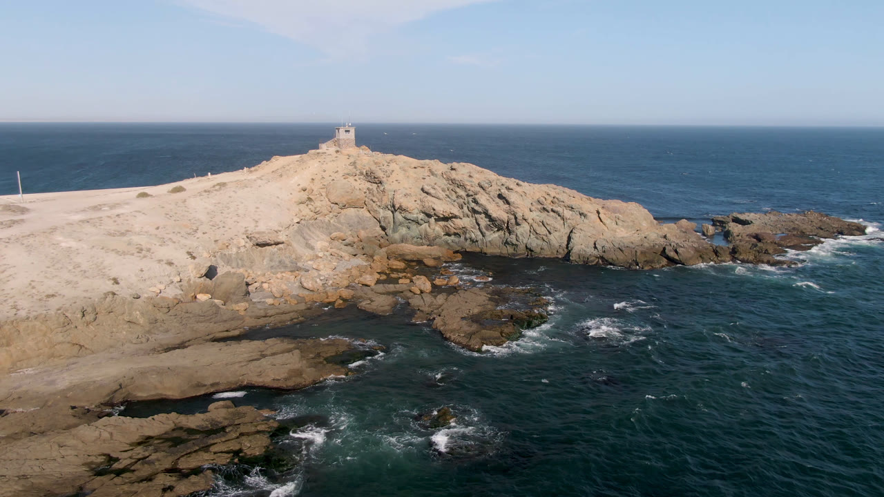 An aerial view frames the tan rocky Bahia Ascuncion headland against the Pacific horizon, with white water breaking around foreground rocks and no visible people.