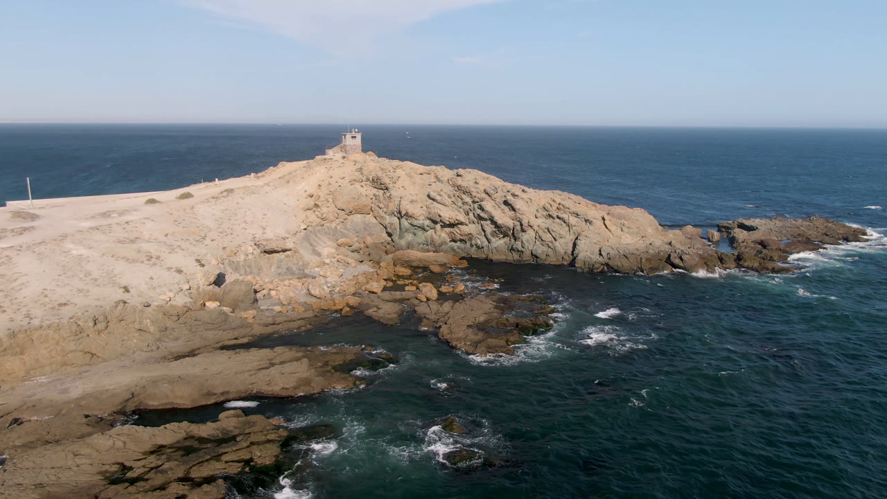 The aerial composition holds the Bahia Ascuncion rocky point in the center, with darker water on the right, surf lines at the base of the rocks, and no visible people.