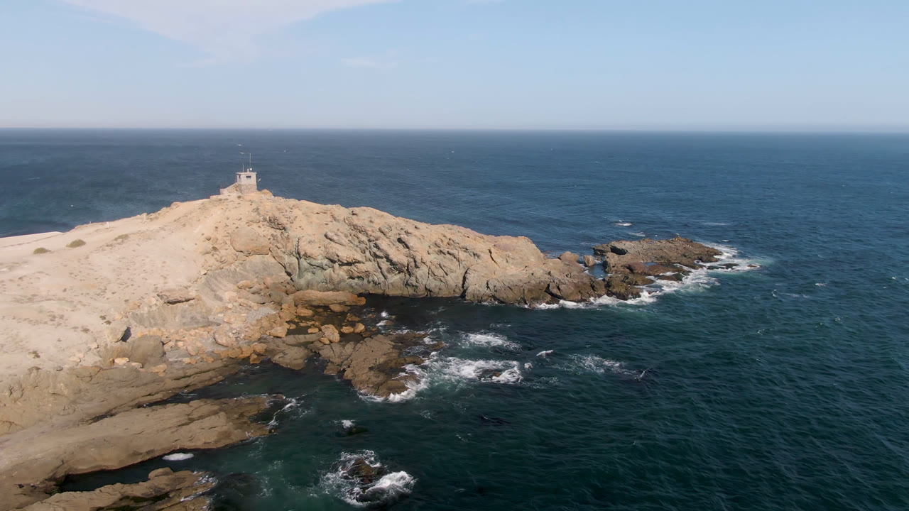 Aerial view in Bahia Ascuncion, Baja California Sur, Mexico, showing a small lighthouse near the left side of a pale rocky headland with dark ocean water and no visible people.