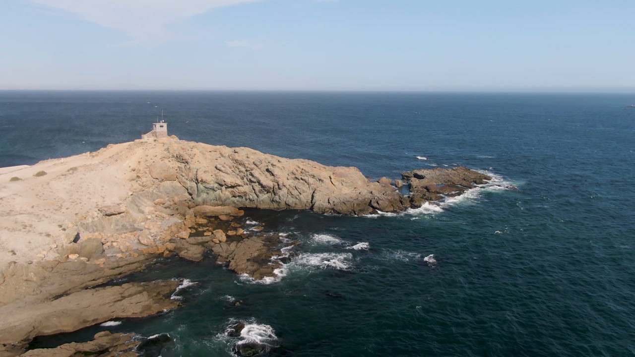 Aerial frame of the Bahia Ascuncion lighthouse sitting higher on the rocky point as white waves break against darker rocks below, with no visible people.