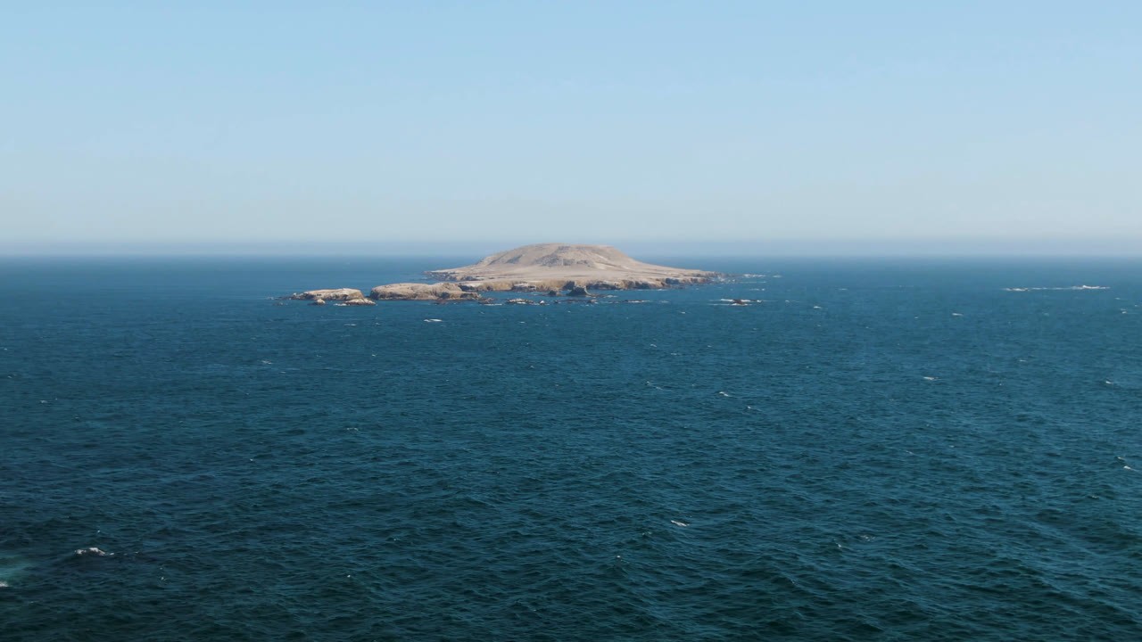 A centered rocky island sits on the horizon above rippled blue ocean near Bahia Ascuncion, with small white wave marks and no visible people.