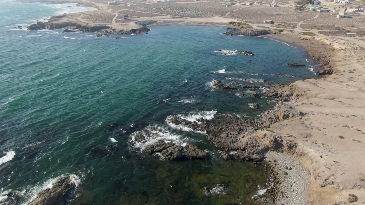 A second aerial view of Bahia Ascuncion shows clear green-blue water, foam around foreground rocks, a sandy right shoreline, scattered scrub inland, and no visible people.