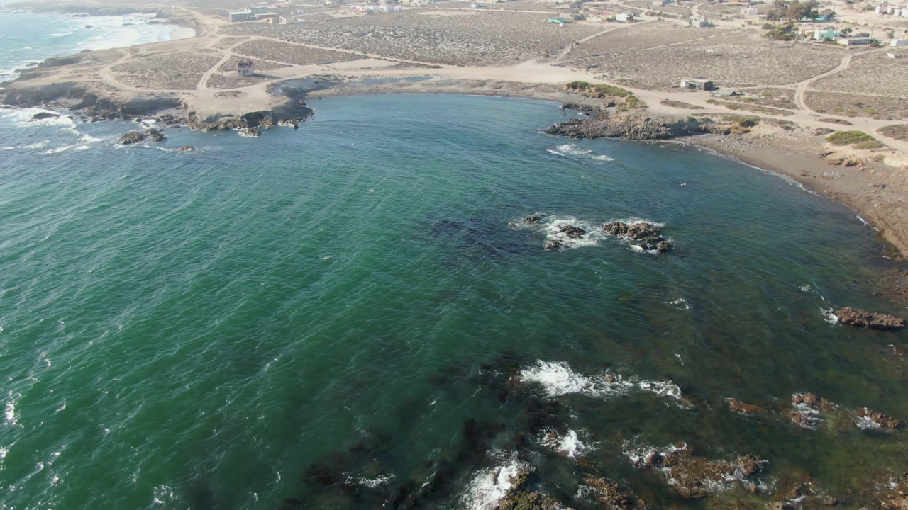 Aerial frame over Bahia Ascuncion showing green-blue water, dark offshore rocks with white foam, a curved sandy beach, pale desert ground, and no visible people.
