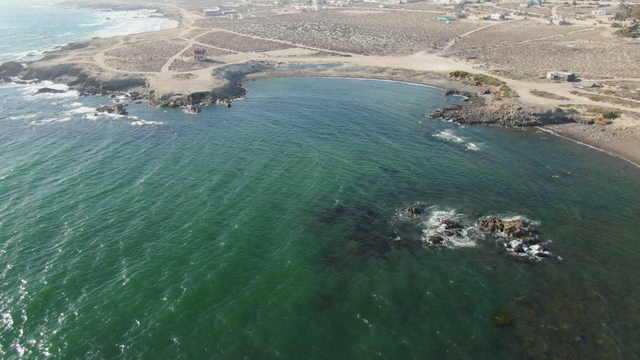 Drone-height frame over Bahia Ascuncion showing open green water across the lower frame, white surf along a rocky inlet, sandy tracks beyond the beach, and no visible people.