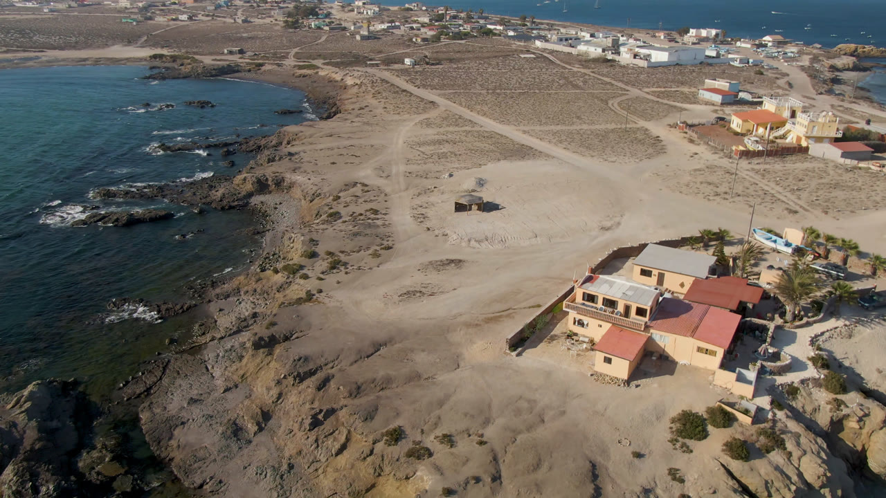 Aerial view of sandy coast, dark rocks, ocean coves, and a few coastal buildings.