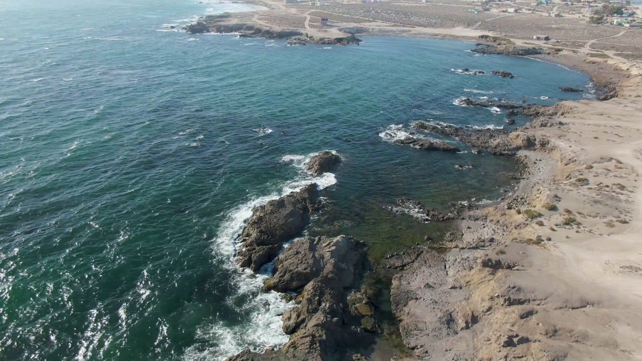 Aerial frame over Bahia Ascuncion with blue-green water, white foam around a large dark rock, pale sandy beach to the right, and no visible people.