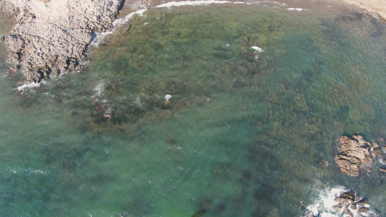 Aerial view over Bahia Ascuncion showing shallow green water around a central rock outcrop, beach at the top edge, and no visible people.