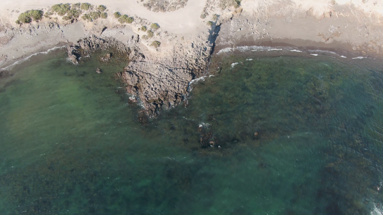 Overhead view of Bahia Ascuncion shoreline with pale sand, rocky outcrops, green shallow water, small surf lines, and no visible people.