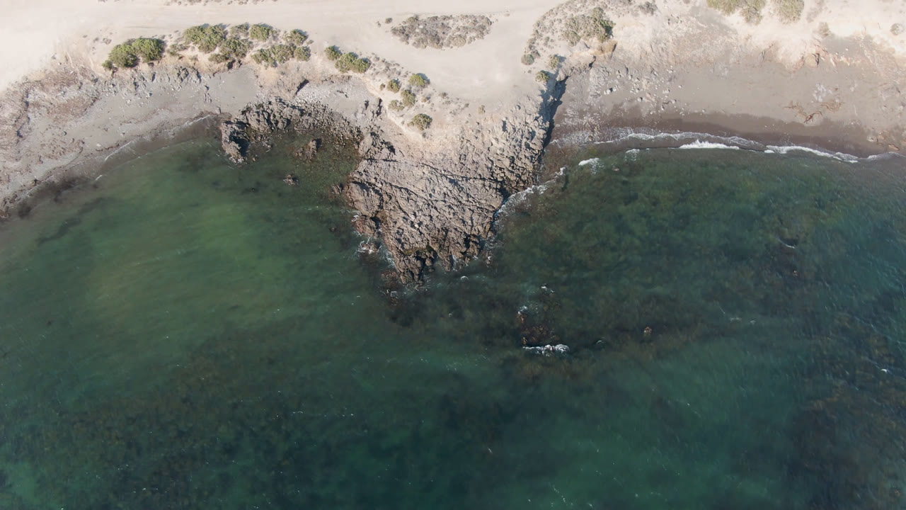 Aerial frame over Bahia Ascuncion showing a central rock point, pale beach above it, translucent green water below, faint wave foam, and no visible people.