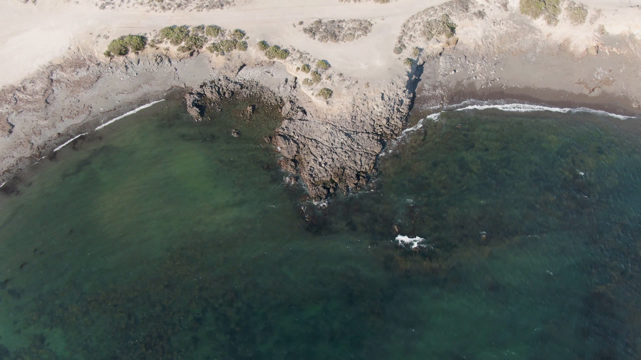 Top-down Bahia Ascuncion view with dunes and scrub near the sand, a jagged rock edge at mid-frame, darker water to the right, and no visible people.