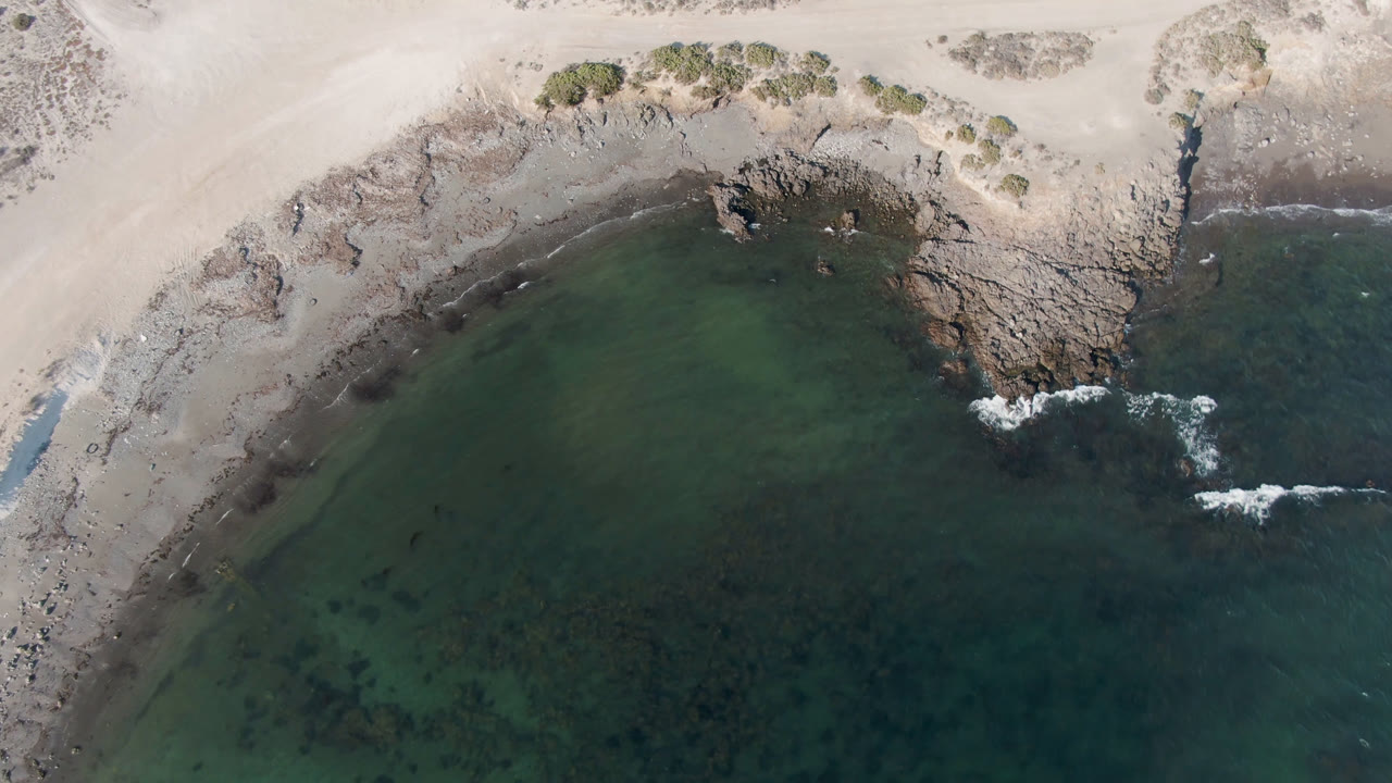 Overhead aerial frame from Bahia Ascuncion shows a pale sandy curve, rocky shoreline, green nearshore water, and no visible people.