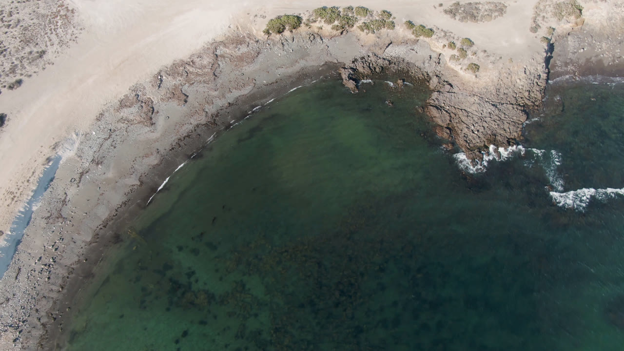 Aerial view over Bahia Ascuncion shows more of the rocky point, pale sand above it, dark green water patches, and no visible people.