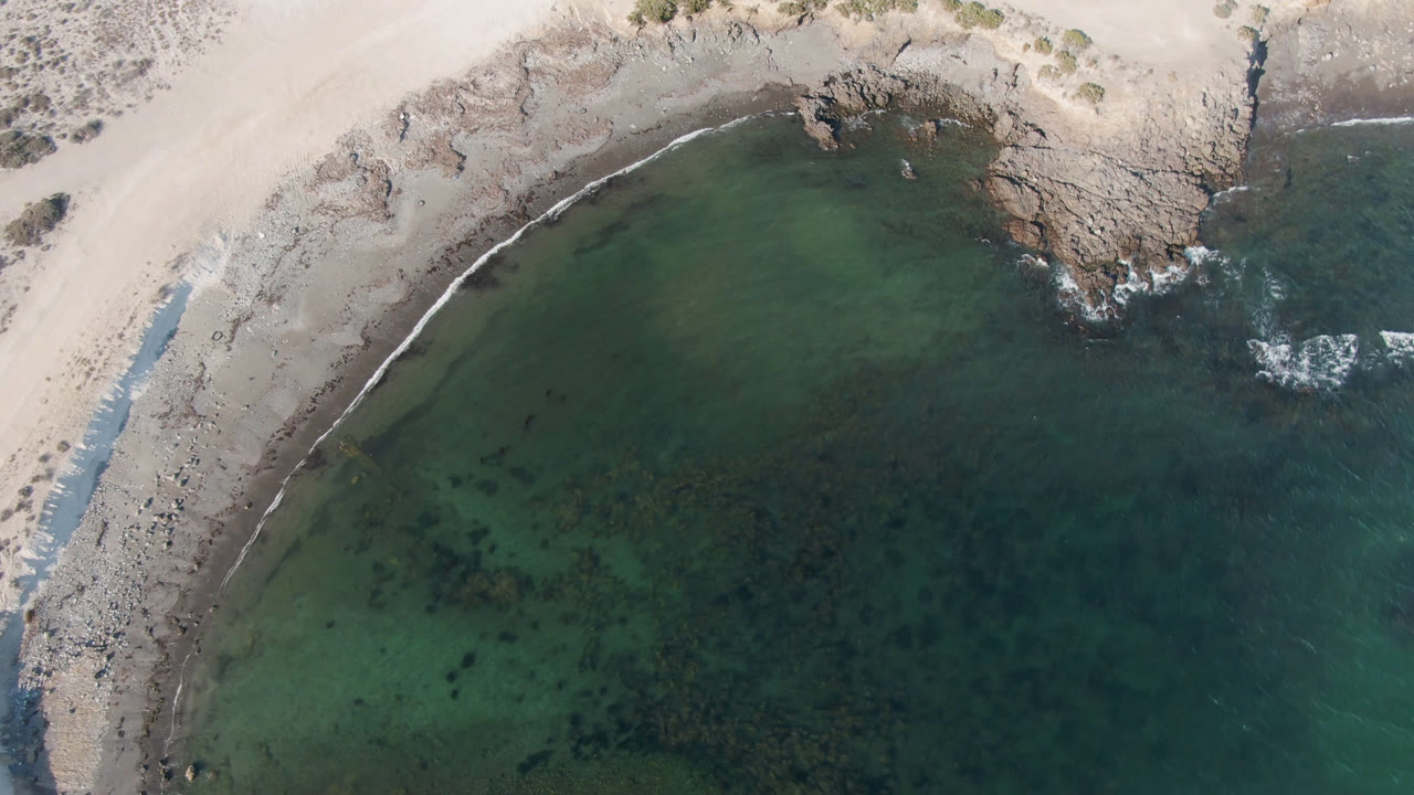 Top-down coastal water beside a sandy and rocky shoreline.