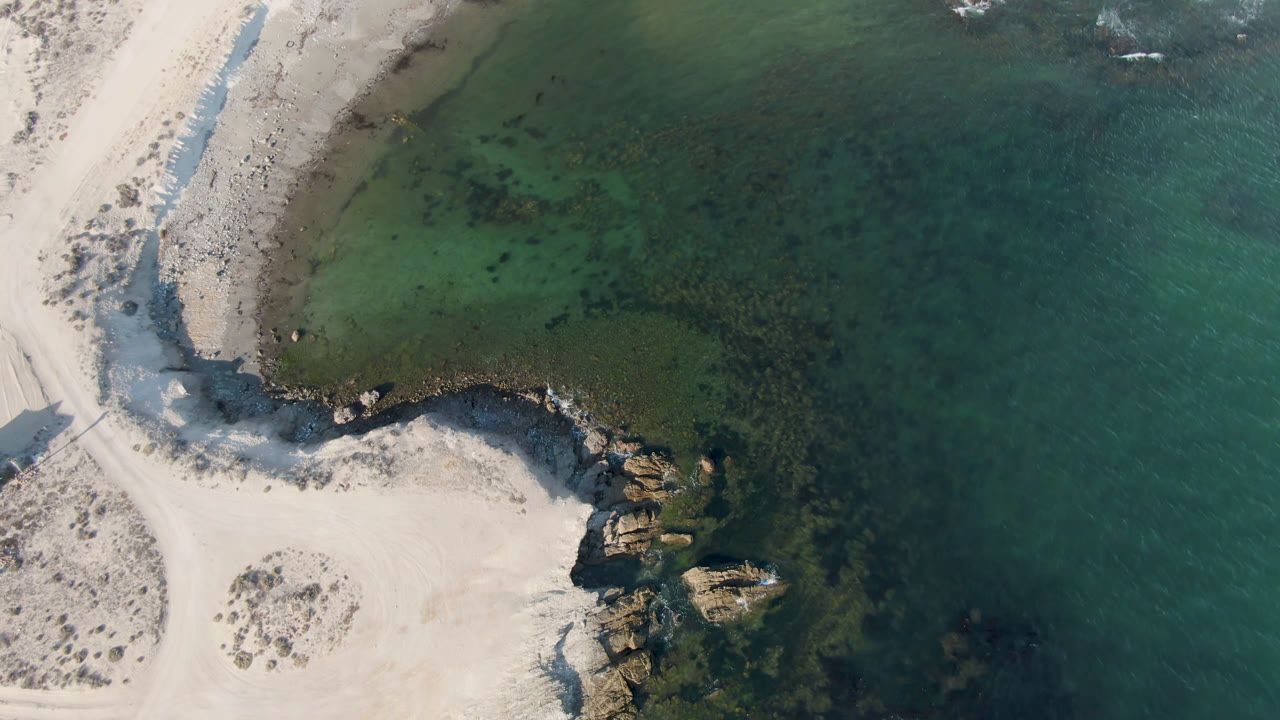 Overhead view of Bahia Ascuncion with pale sand on the left, dark shoreline rocks, clear green water on the right, and no visible people.