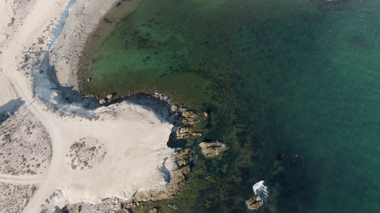 Aerial frame from Bahia Ascuncion showing a wider section of pale beach, scattered scrub on sand, shoreline rocks, green shallows, and no visible people.