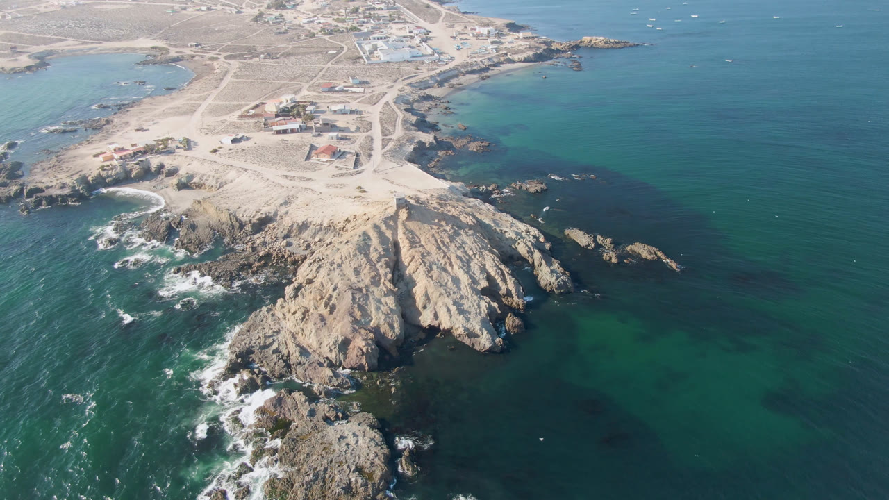 Aerial view over Bahia Ascuncion, Baja California Sur, with jagged rocks in the foreground, sandy tracks and small buildings inland, turquoise ocean around the point, and no visible people.