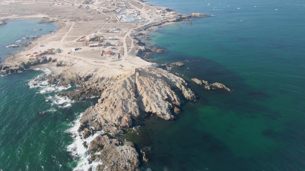 Opening aerial frame over Bahia Ascuncion, Baja California Sur, with a rocky point below, teal water to the right, pale sand tracks inland, scattered buildings near shore, and no visible people.