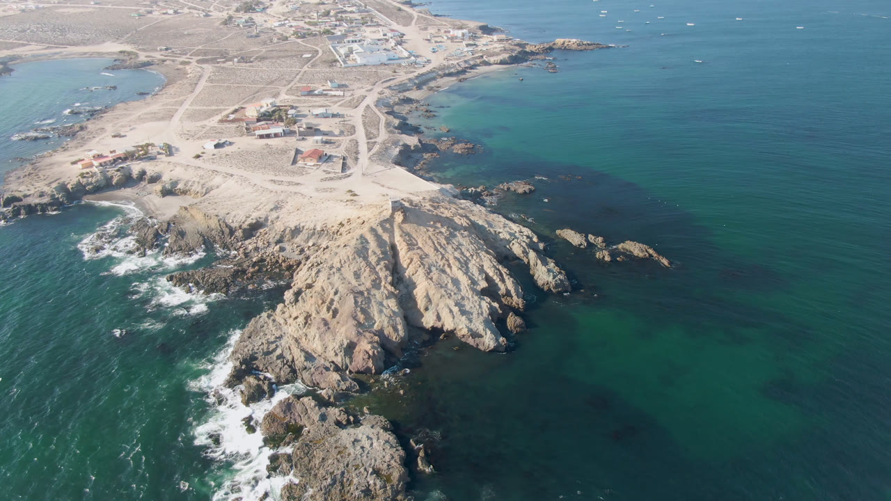 Aerial frame over Bahia Ascuncion showing the rocky headland centered lower in view, white waves on the left edge, turquoise shallows near shore, inland roads, small buildings, and no visible people.
