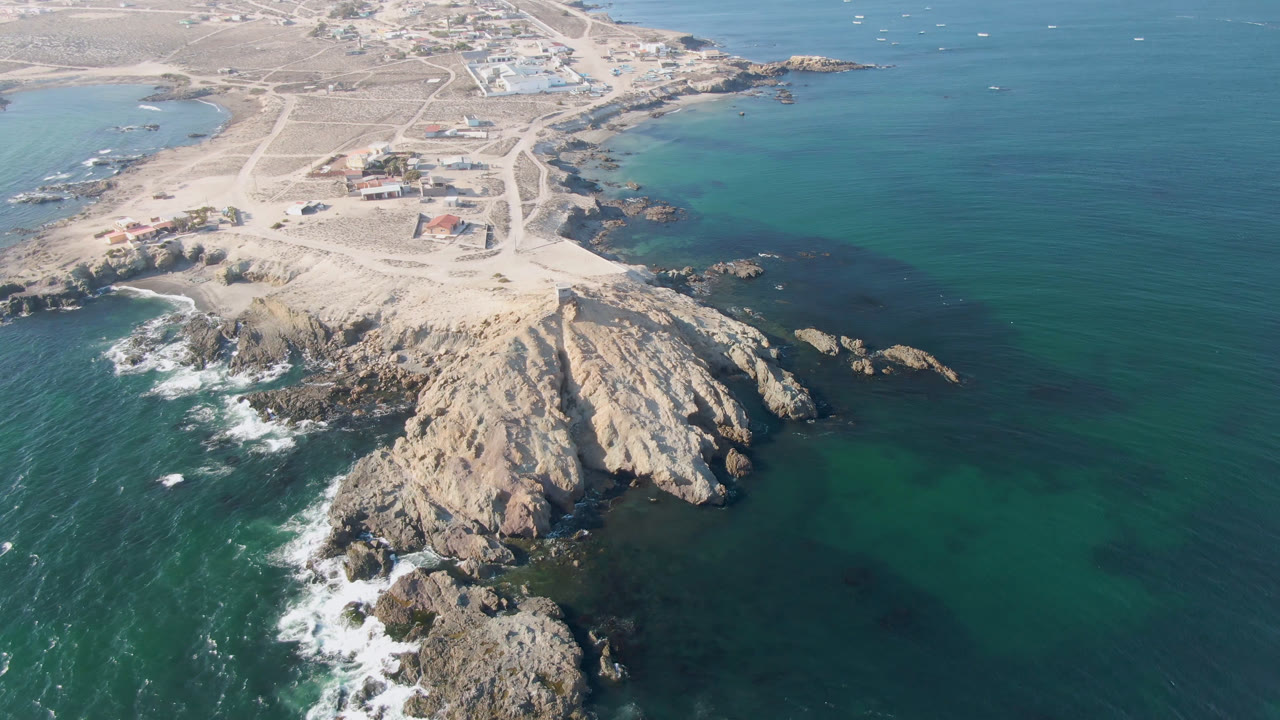 Aerial coastal frame with rocks, open ocean, sandy roads, and distant shore buildings