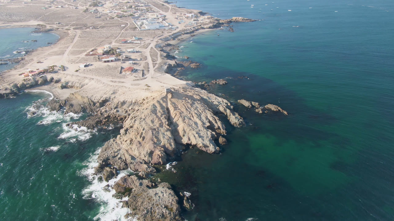 Aerial frame over Bahia Ascuncion, Baja California Sur, showing pale rocky headland, turquoise water, white surf, sandy roads, low buildings, and no visible people.