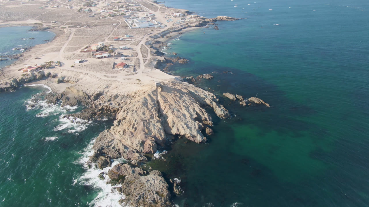 Second aerial frame from Bahia Ascuncion with jagged rocks centered lower in the image, blue-green ocean on both sides, white waves at the left edge, and no visible people.