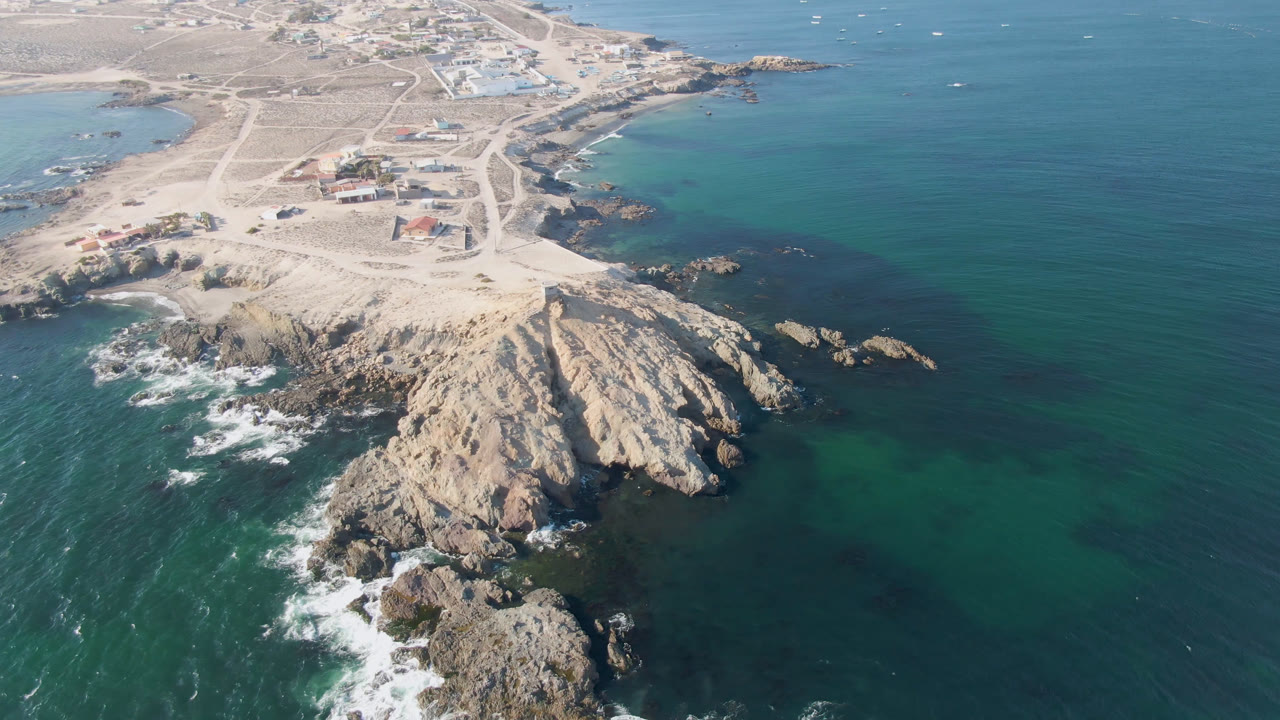 High aerial angle on rocky point, surf, open water, and coastal settlement.