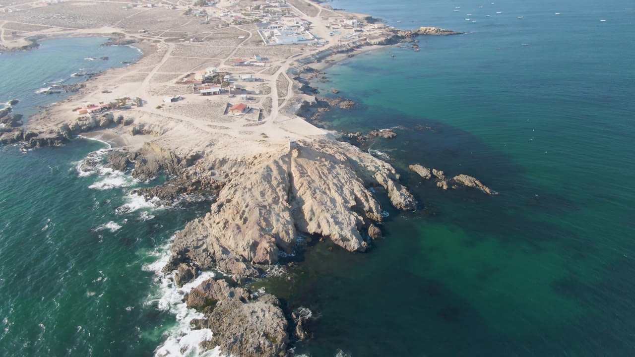 Aerial frame over Bahia Ascuncion, Baja California Sur, showing a rocky point in the foreground, turquoise water on both sides, pale sand roads inland, and no visible people.