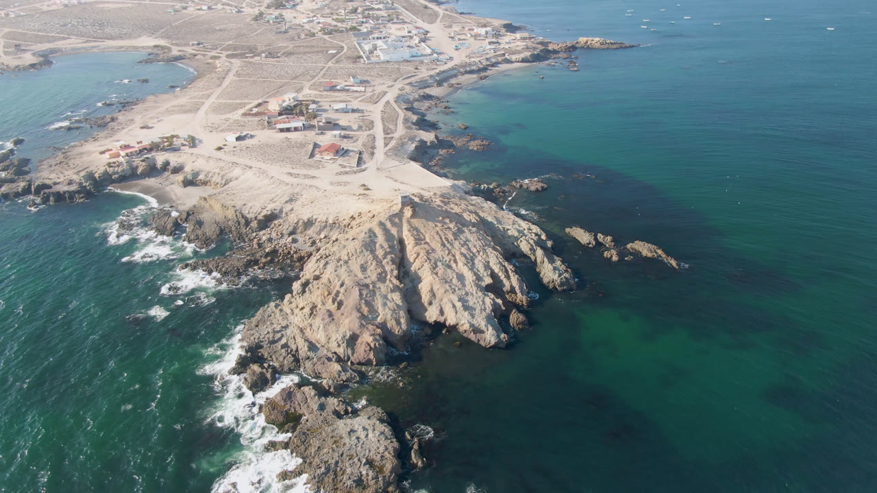Drone-height view of Bahia Ascuncion with white waves breaking along the left side of the rocky point and darker teal water wrapping around offshore rocks, with no visible people.