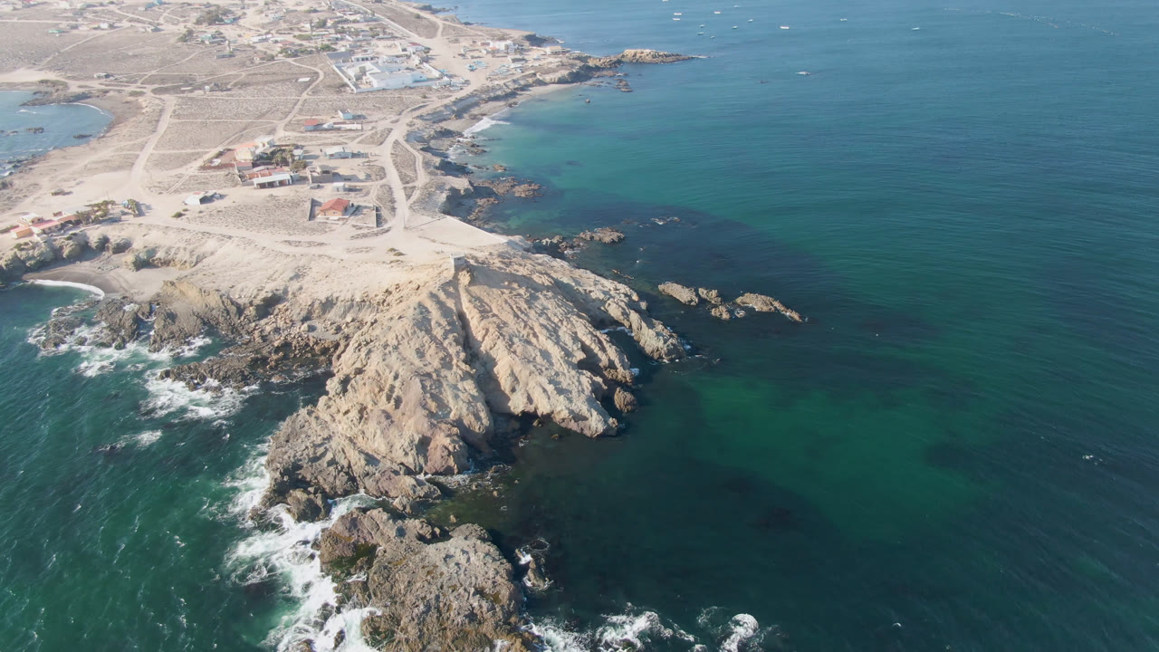Aerial frame over Bahia Ascuncion showing pale rock formations jutting into turquoise and deep blue ocean, white surf, sandy tracks, scattered buildings, and no visible people.