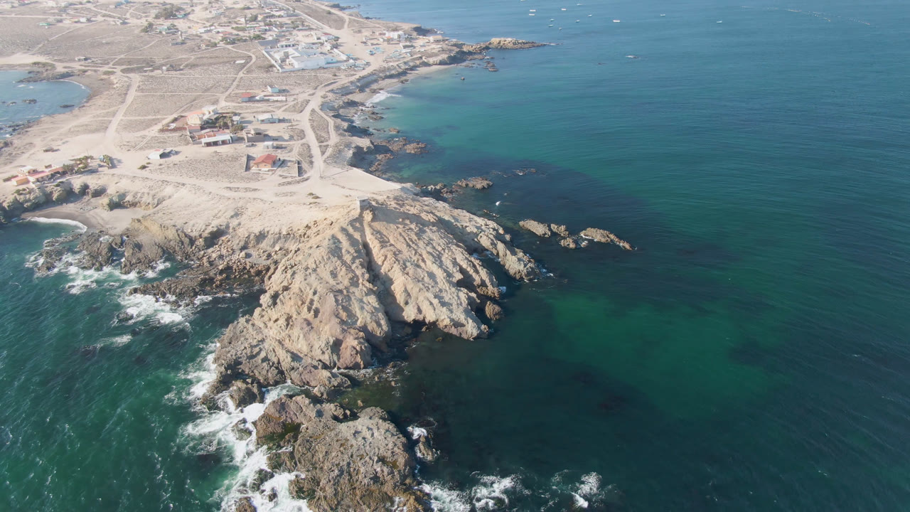 High aerial view in Bahia Ascuncion with the rocky point centered lower in frame, green shallows around the rocks, open blue water to the right, distant buildings, and no visible people.