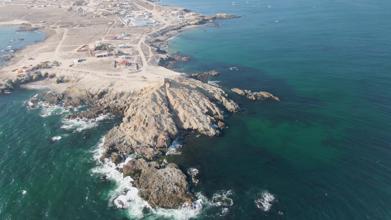 Aerial frame over Bahia Ascuncion showing a tan rocky headland, sandy beach behind it, green-blue ocean, white surf around foreground rocks, and no visible people.