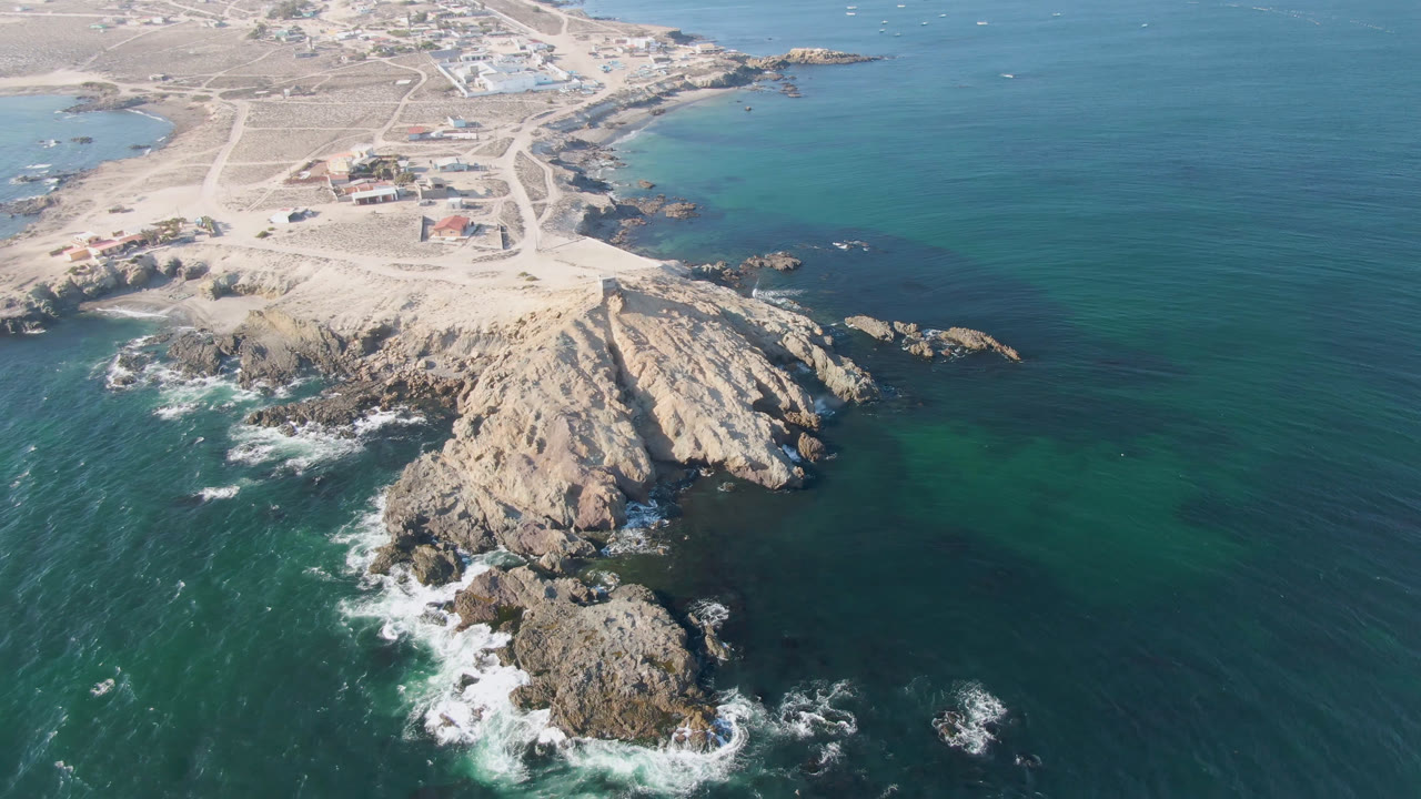 Aerial frame over Bahia Ascuncion with the rocky point centered lower in the view, surf curling around the rocks, pale beach and roads above, and no visible people.