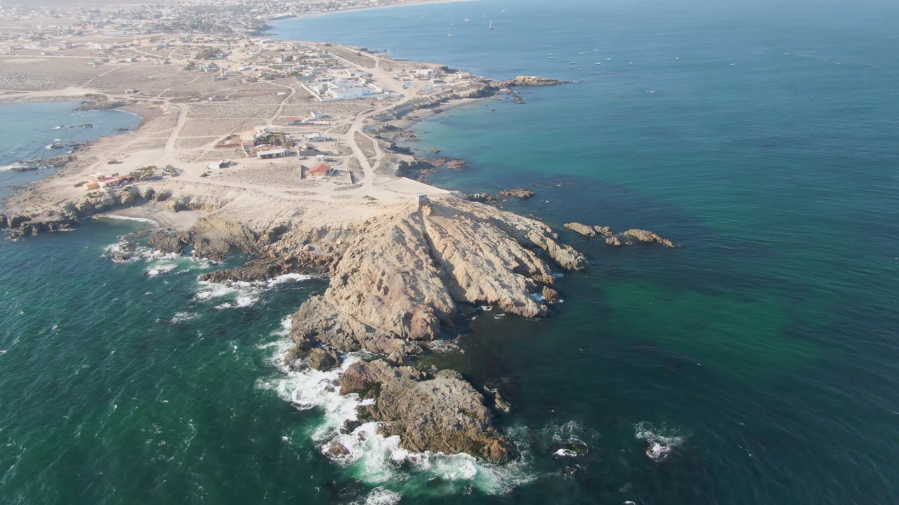 High aerial frame over Bahia Ascuncion, Baja California Sur, with a rocky point in the foreground, pale roads and scattered buildings inland, teal ocean on both sides, and no visible people.