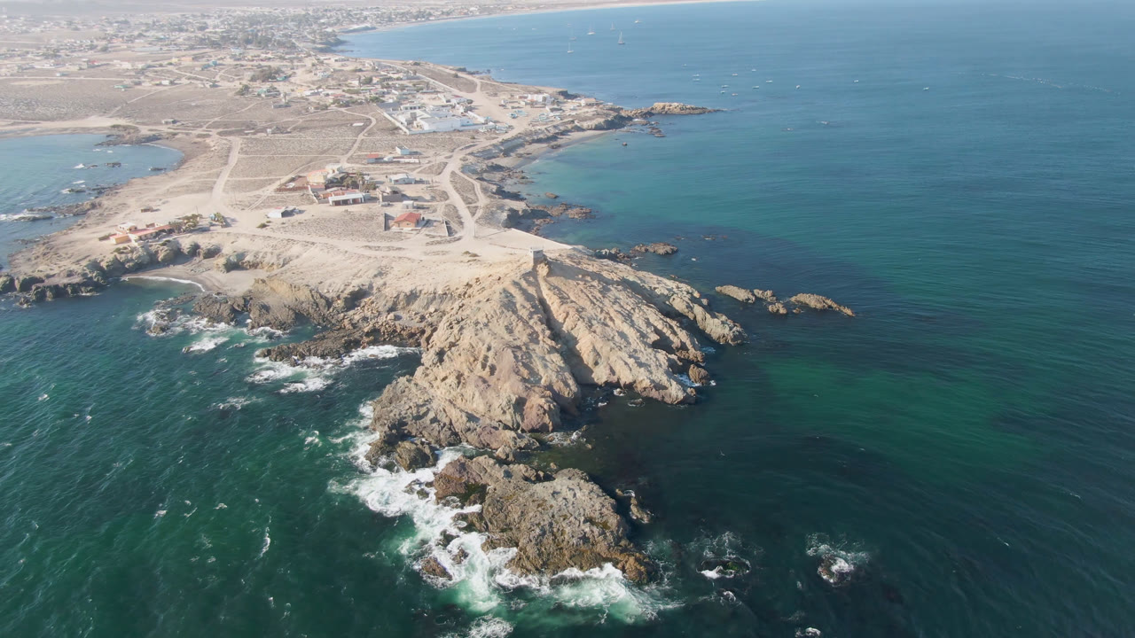 Drone frame over Bahia Ascuncion showing white surf around the lower rocks, turquoise shallows near shore, open blue water to the right, inland roads, and no visible people.