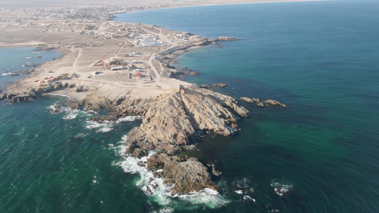 Wide drone frame of a rocky headland with ocean and a pale coastal village area behind it.