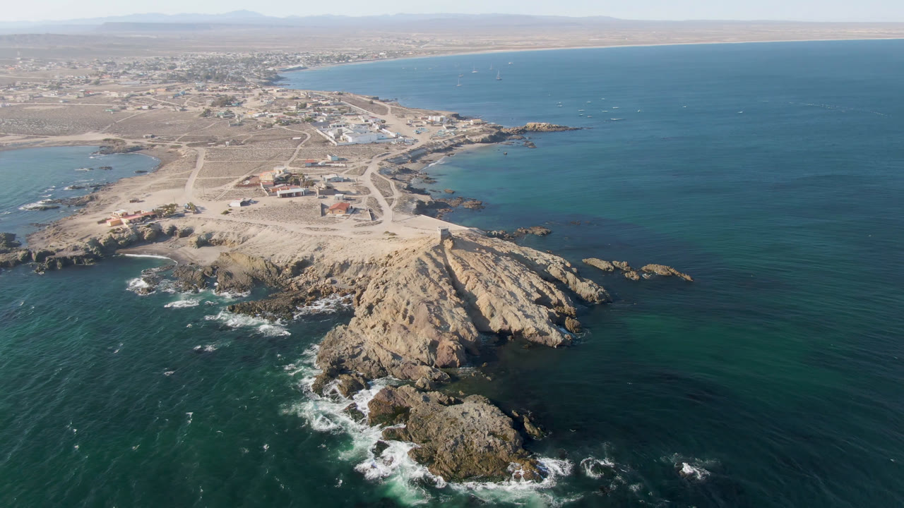 Aerial frame over Bahia Ascuncion, Baja California Sur, Mexico showing a rocky point in the foreground, teal ocean on both sides, sandy roads, low buildings, distant hills, and no visible people.