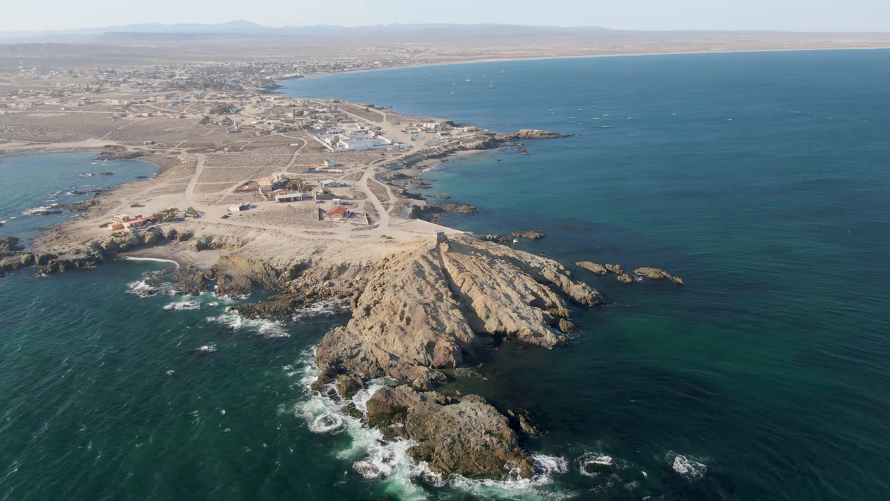 Drone frame over Bahia Ascuncion with white surf breaking around foreground rocks, a pale sandy point, scattered low buildings behind it, open bay water to the right, and no visible people.