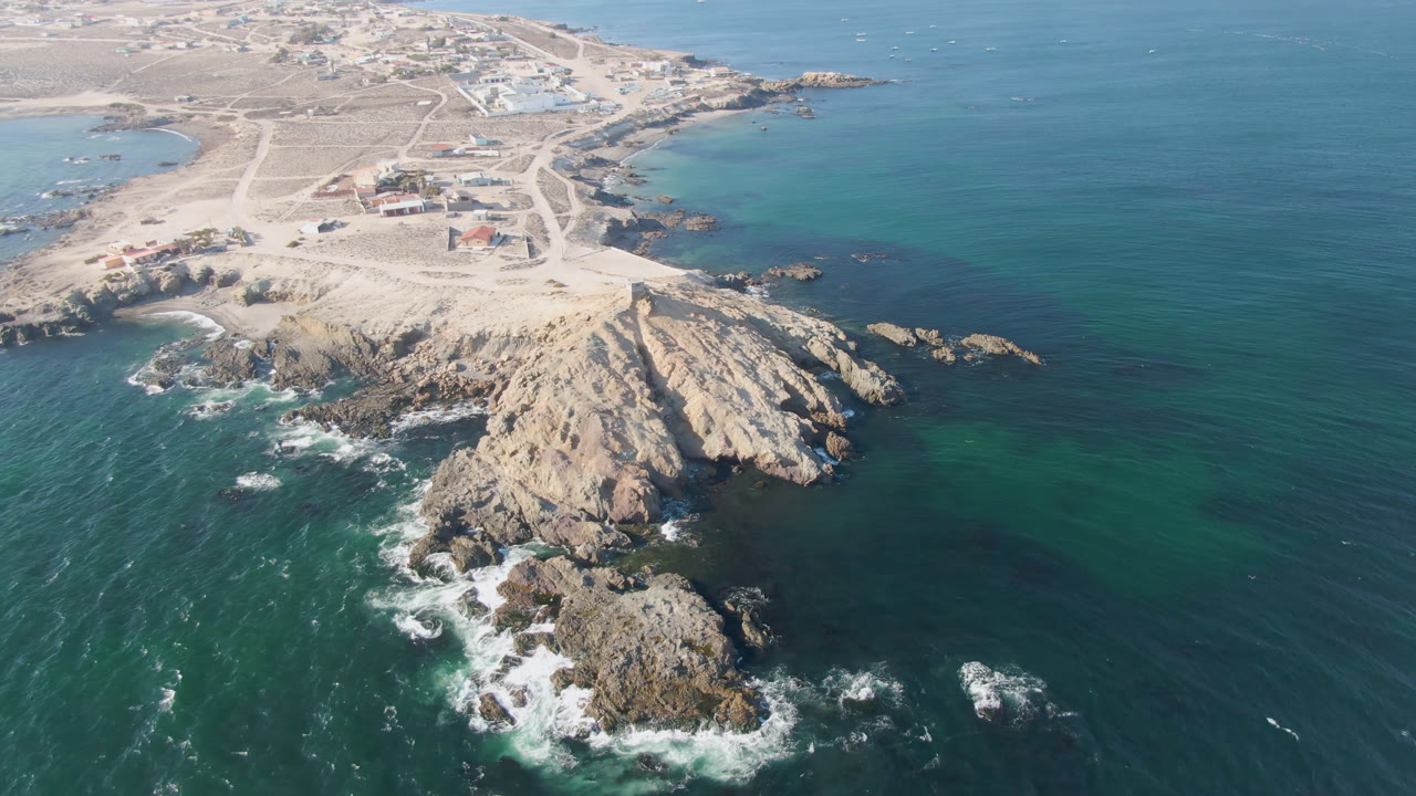 High aerial view over Bahia Ascuncion, Baja California Sur, showing a pale rocky headland, offshore rocks, turquoise water, white surf, sandy tracks, low buildings, and no visible people.