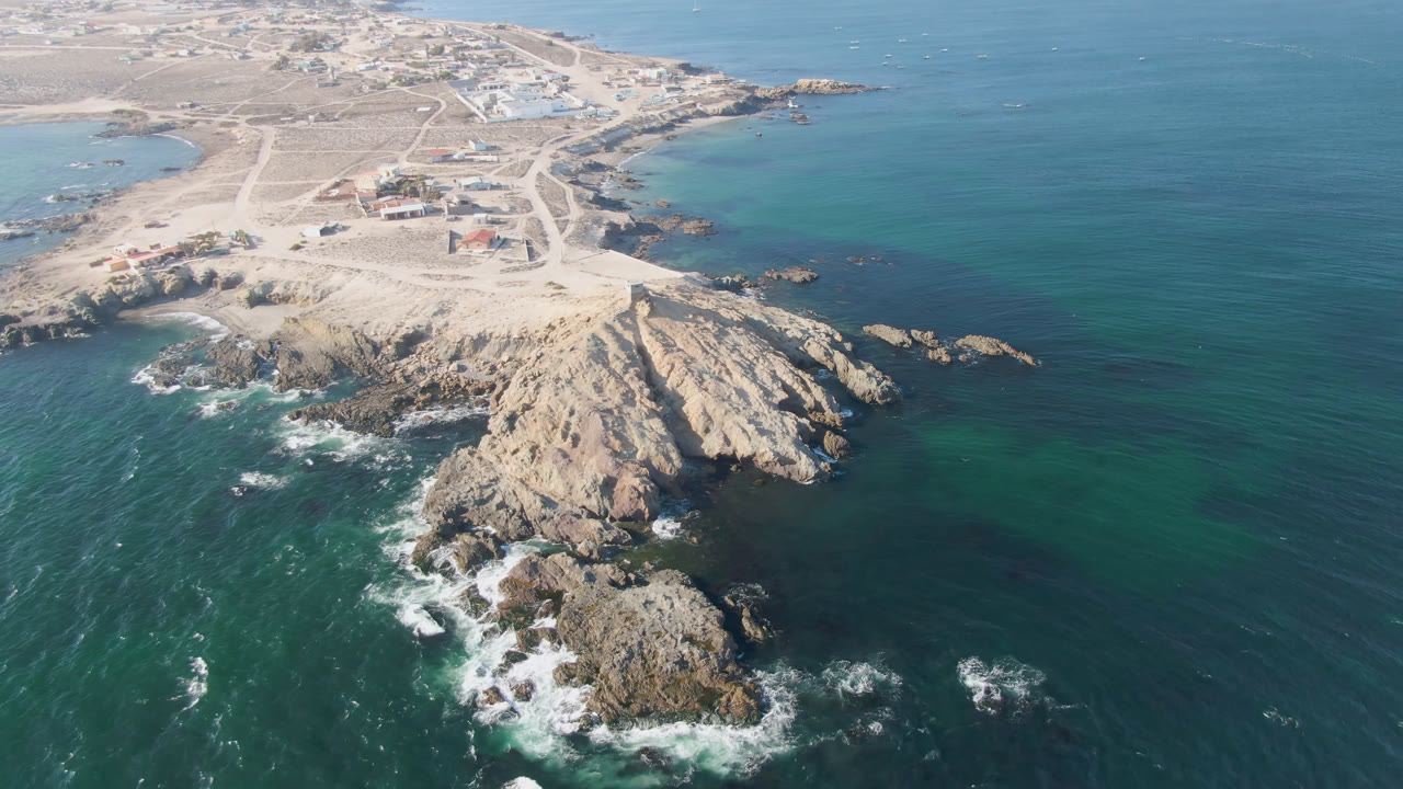 Aerial frame over Bahia Ascuncion with the rocky point centered lower in frame, white water breaking along the left edge, clear blue-green shallows, inland roads, buildings, and no visible people.