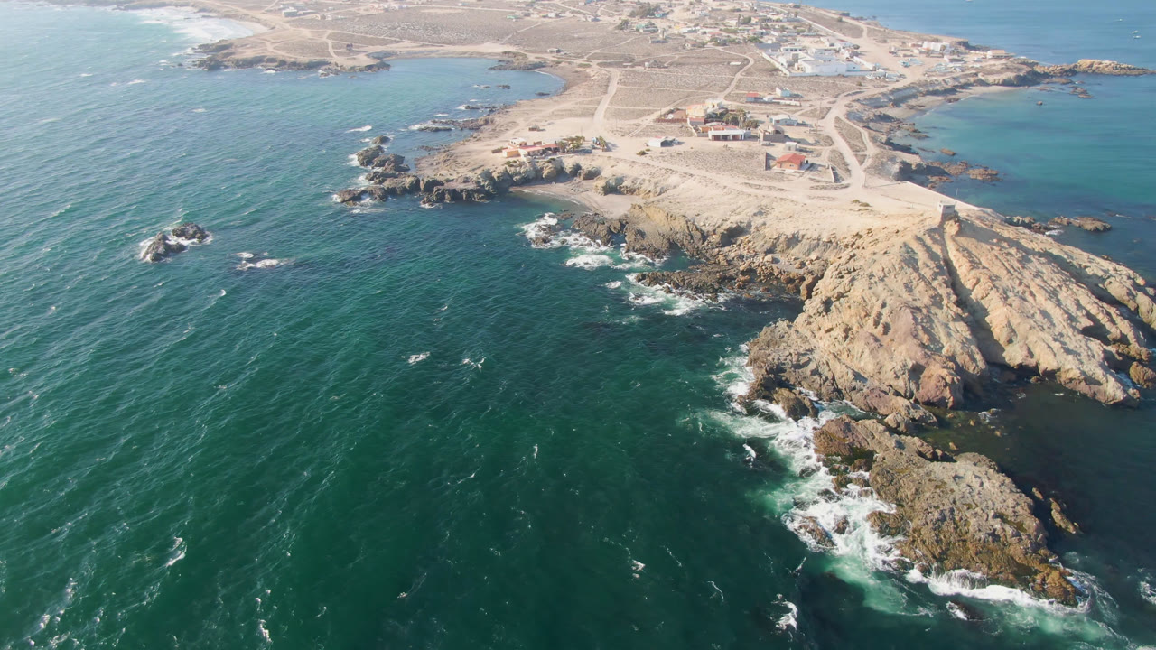 Aerial frame over Bahia Ascuncion, Baja California Sur, showing teal water around tan rock formations, sandy tracks, distant low buildings, and no visible people.