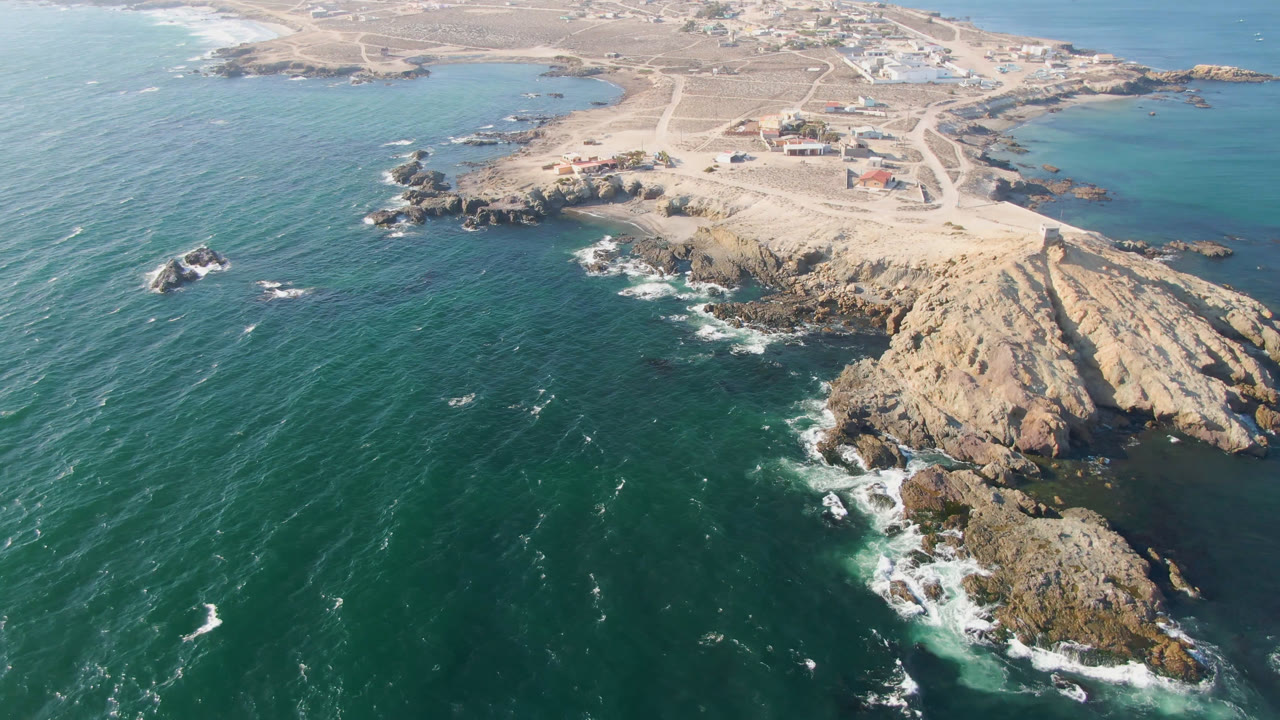 Aerial view in Bahia Ascuncion with white surf wrapping the lower rock outcrops, open green-blue water to the left, sandy roads above, and no visible people.
