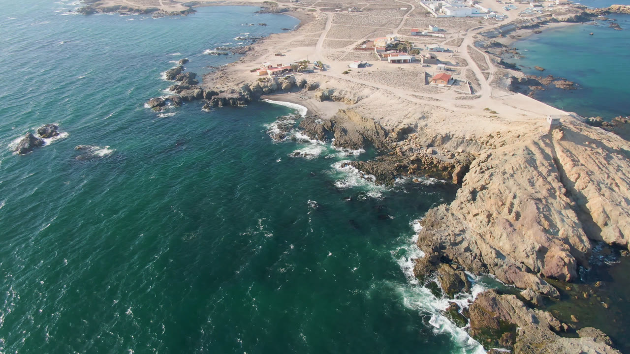 Aerial view of rocky coastline, teal ocean, sandy roads, and small buildings.