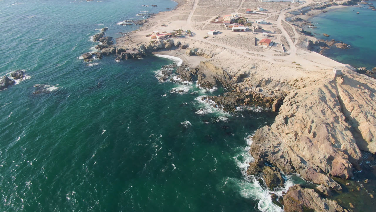 Second aerial frame in Bahia Ascuncion with more open water at left, waves breaking against dark rocks below the pale bluff, sandy roads, small buildings, and no visible people.